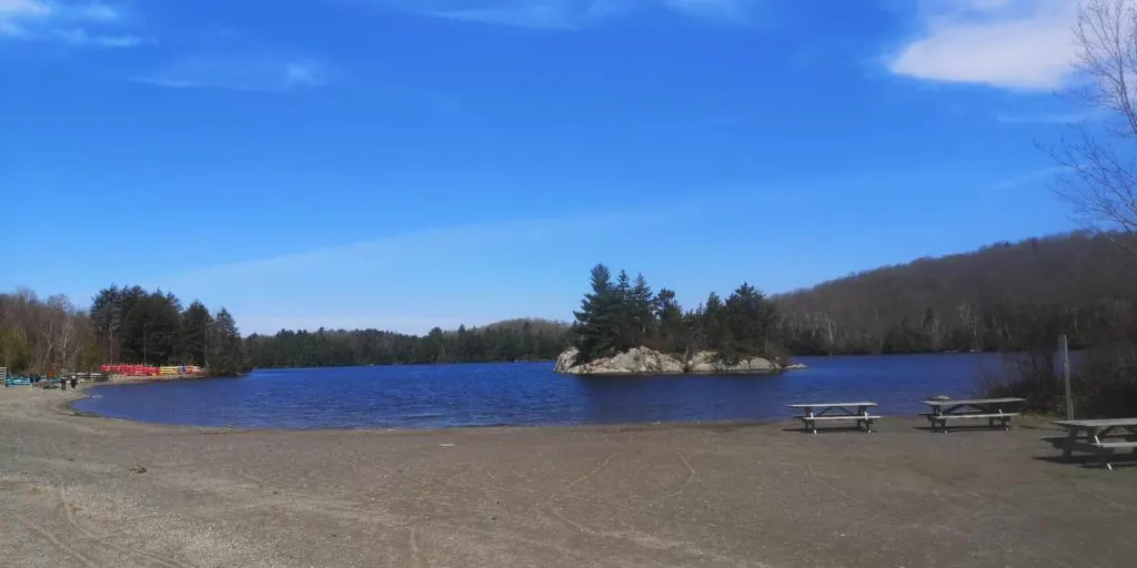 Lac au Mont Orford avec un îlot rocheux, une plage de sable et des tables de pique-nique